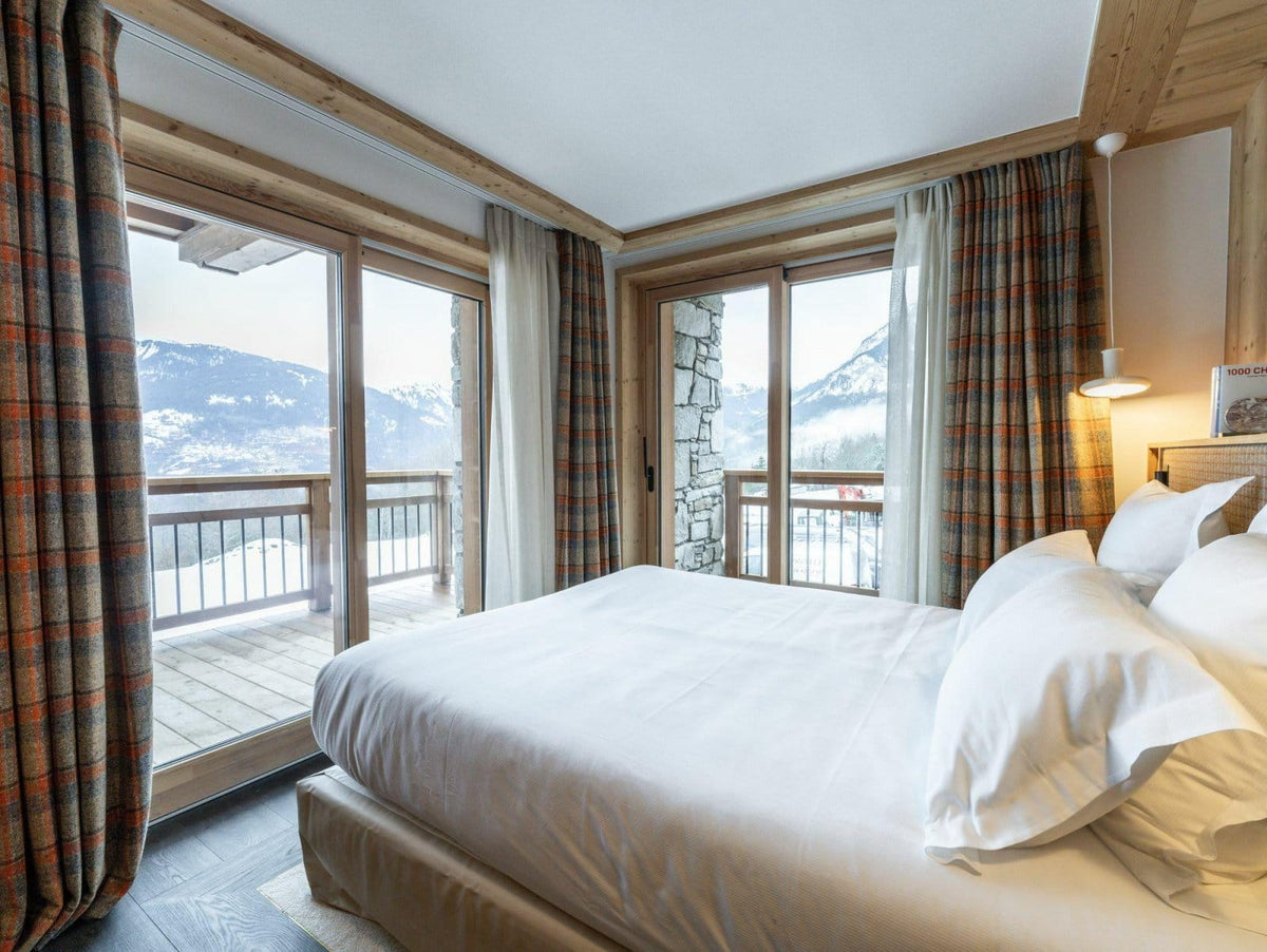 A luxury ski chalet bedroom featuring a large bed with crisp white linens, rustic wooden ceiling beams, and floor-to-ceiling windows with plaid curtains, offering a stunning view of snow-covered mountains.