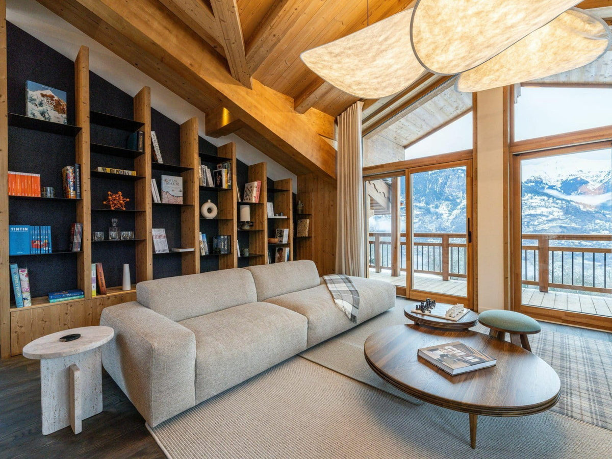 A modern light beige fabric sofa in a rustic wooden chalet living room, with a round wooden coffee table, built-in wooden bookshelves, and large windows overlooking a snowy mountain landscape.