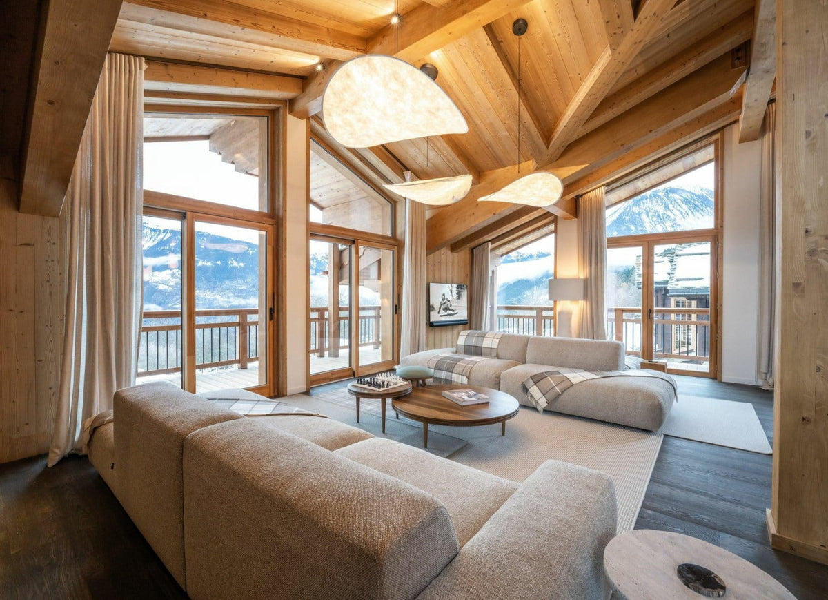 A modern mountain chalet living room featuring a large beige sectional sofa, round wooden coffee tables, and large windows with views of snowy mountains. The room has a high wood-beam ceiling and warm ambient lighting.