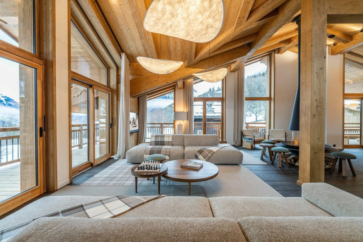 A modern mountain chalet living room with a large beige sectional sofa, round wooden coffee tables, exposed wood ceiling beams, and floor-to-ceiling windows looking out to snowy mountain views.