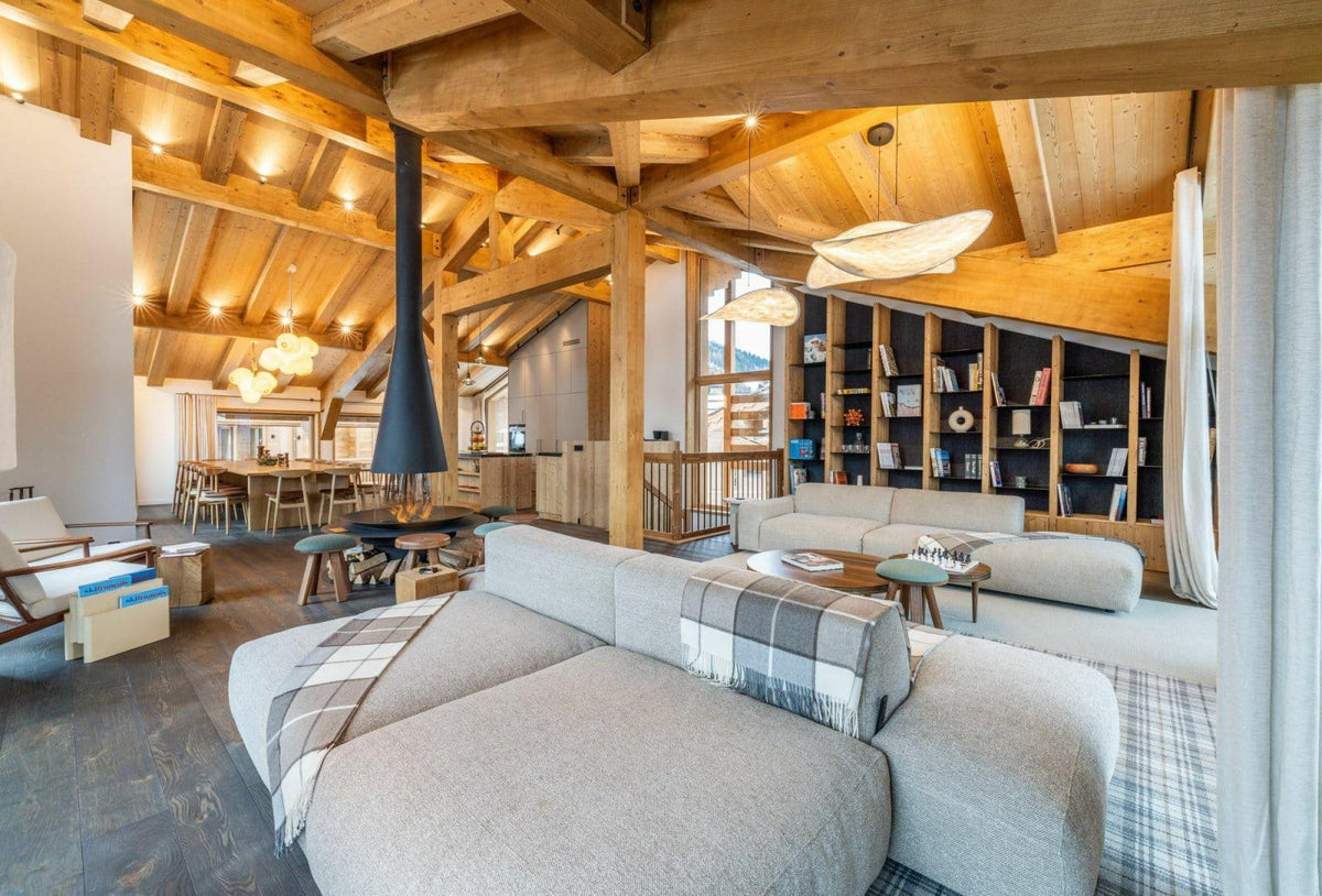 A modern light gray sectional sofa in a rustic, wood-beamed chalet living room with a black fireplace chimney, wooden ceiling beams, and built-in bookshelves.
