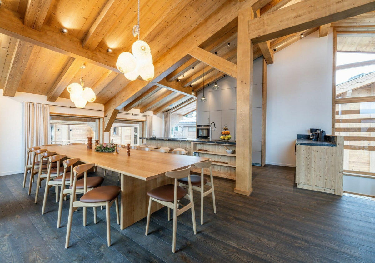 A modern alpine-style dining room with a large wooden dining table and chairs, set under a high wood-beamed ceiling with pendant lights. A kitchen is visible in the background, and the room has dark wood floors and large windows.