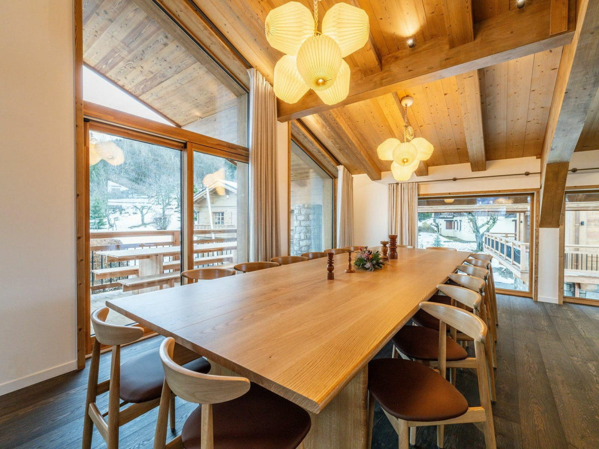 A long wooden dining table with matching wooden chairs in a bright, modern chalet-style dining room with large windows, wood ceiling beams, and hanging pendant lights.