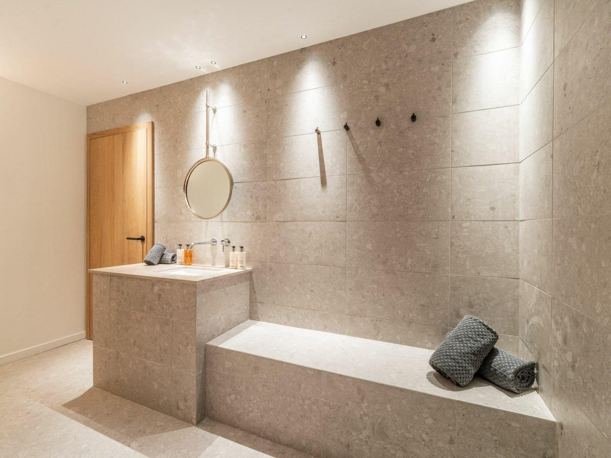 A modern bathroom interior with a stone-tile wall, built-in bench, vanity counter, round wall mirror, and two rolled dark towels on the bench.