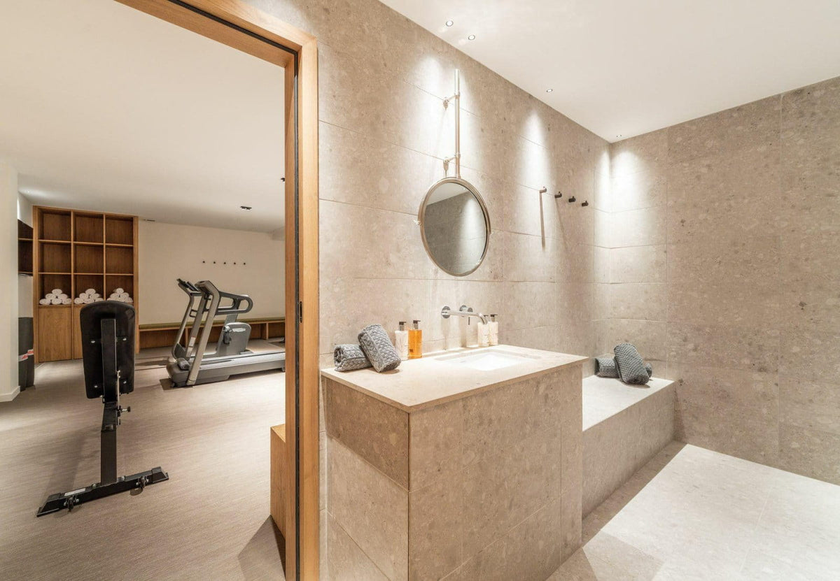 A modern bathroom interior with beige stone walls and floor, featuring a built-in vanity, round wall mirror, towel bars, and a doorway opening into a gym area with a treadmill and exercise equipment.