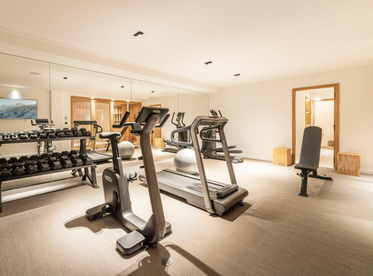 A modern home gym interior with cardio equipment including treadmills and an elliptical machine, plus a weight rack with dumbbells and a bench, set in a spacious room with mirrors and wood flooring.
