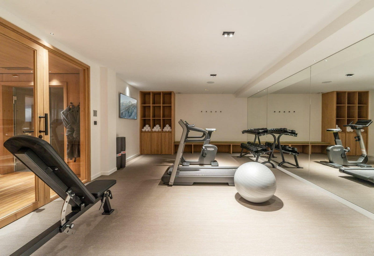 A spacious home gym interior with multiple exercise machines, including a treadmill, elliptical, leg press, and a stability ball, set against beige walls and wood shelving.
