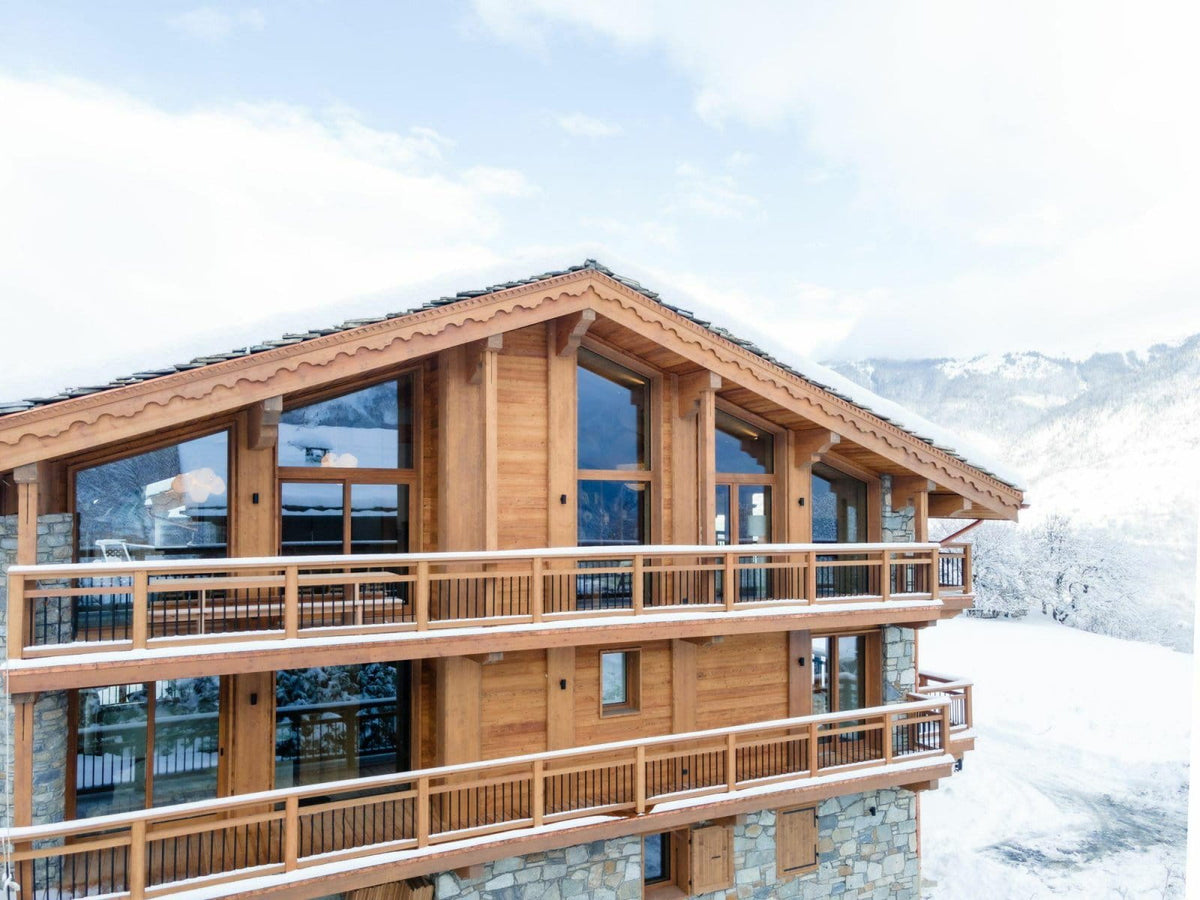 A modern alpine chalet with warm wood paneling, stone foundations, large glass windows, and snow-covered balconies, set against a snowy mountain landscape under a pale sky.
