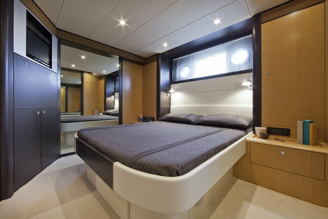 A modern yacht or luxury cabin bedroom featuring a low-profile platform bed with dark gray bedding, sleek dark wood and white cabinetry, and a built-in television screen on the left.