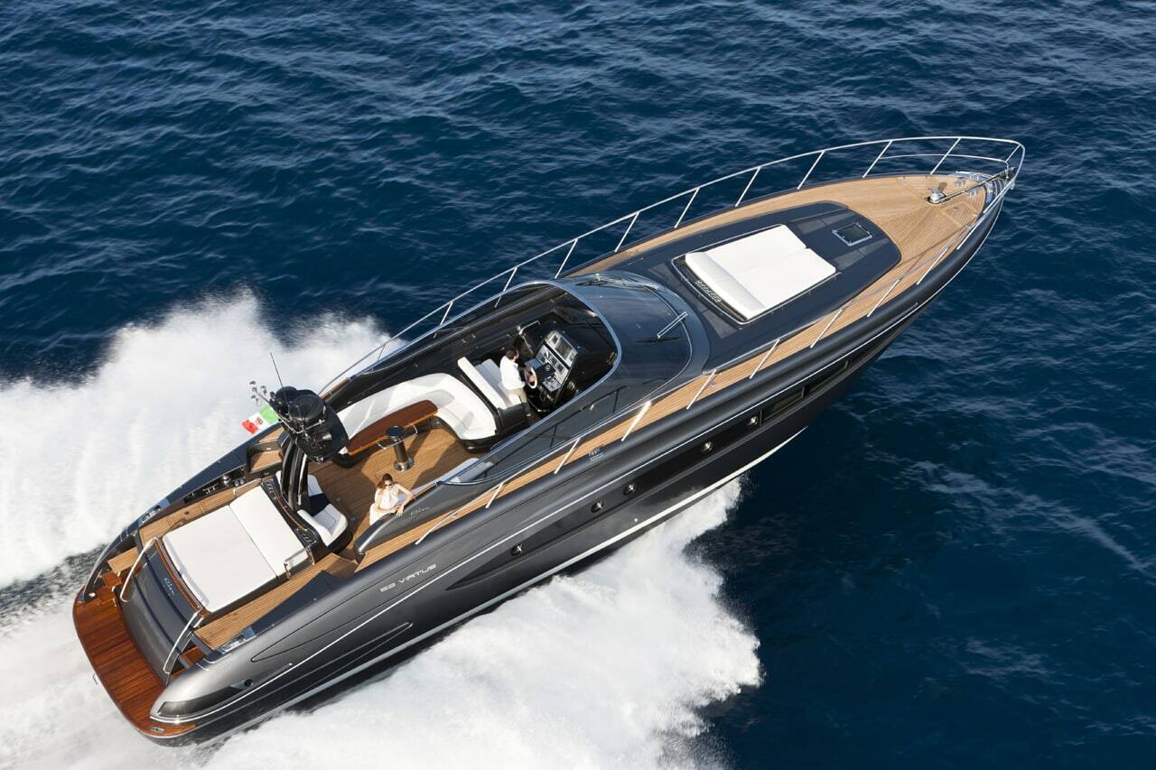 A luxury motor yacht with a black hull and tan wood-look trim, captured from an aerial perspective as it cuts through deep blue ocean water and creates a white wake.