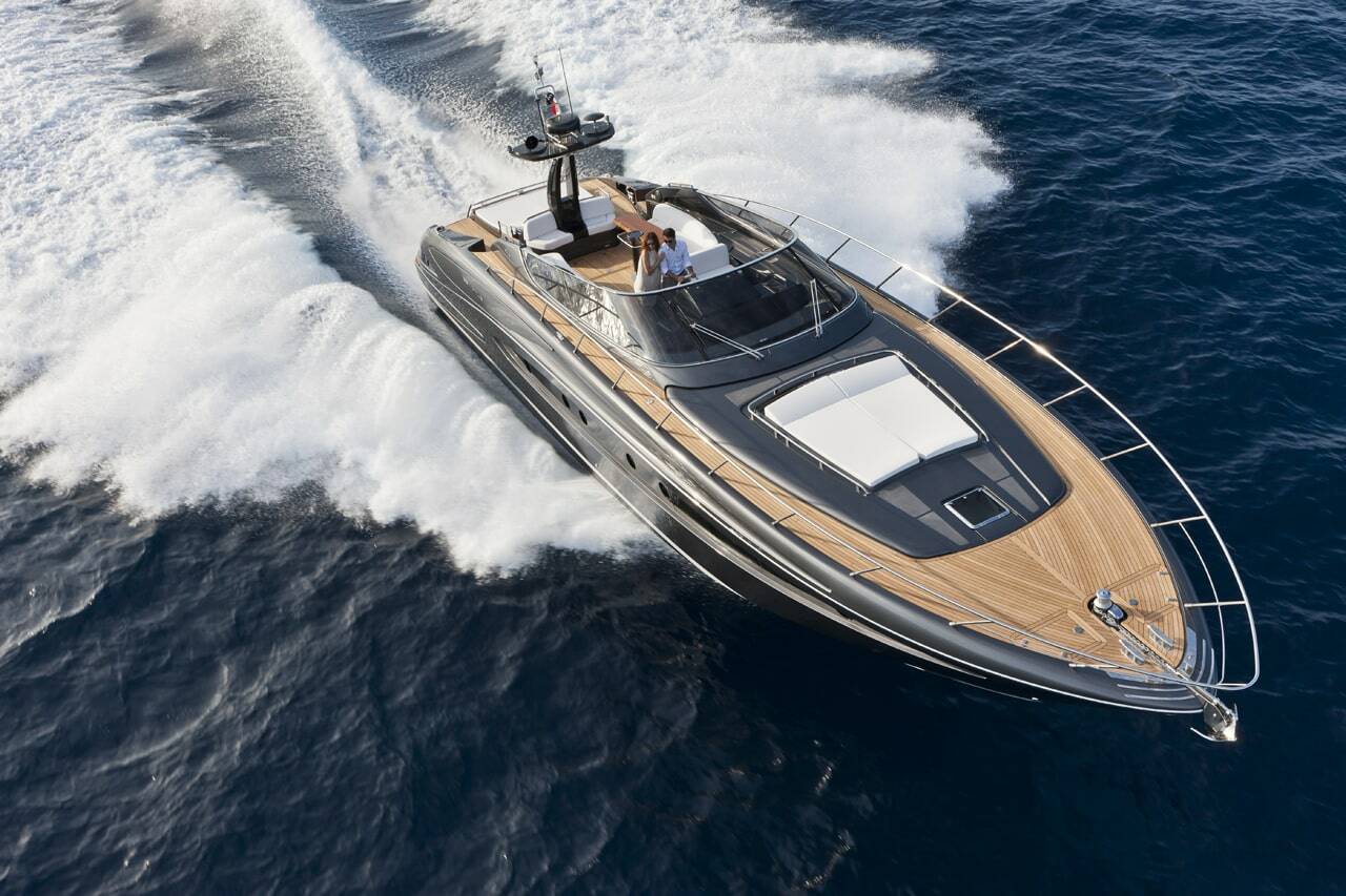 A luxury motor yacht with a dark gray exterior and light wood deck flooring, captured from an aerial perspective as it cuts through deep blue ocean water, creating a white wake.