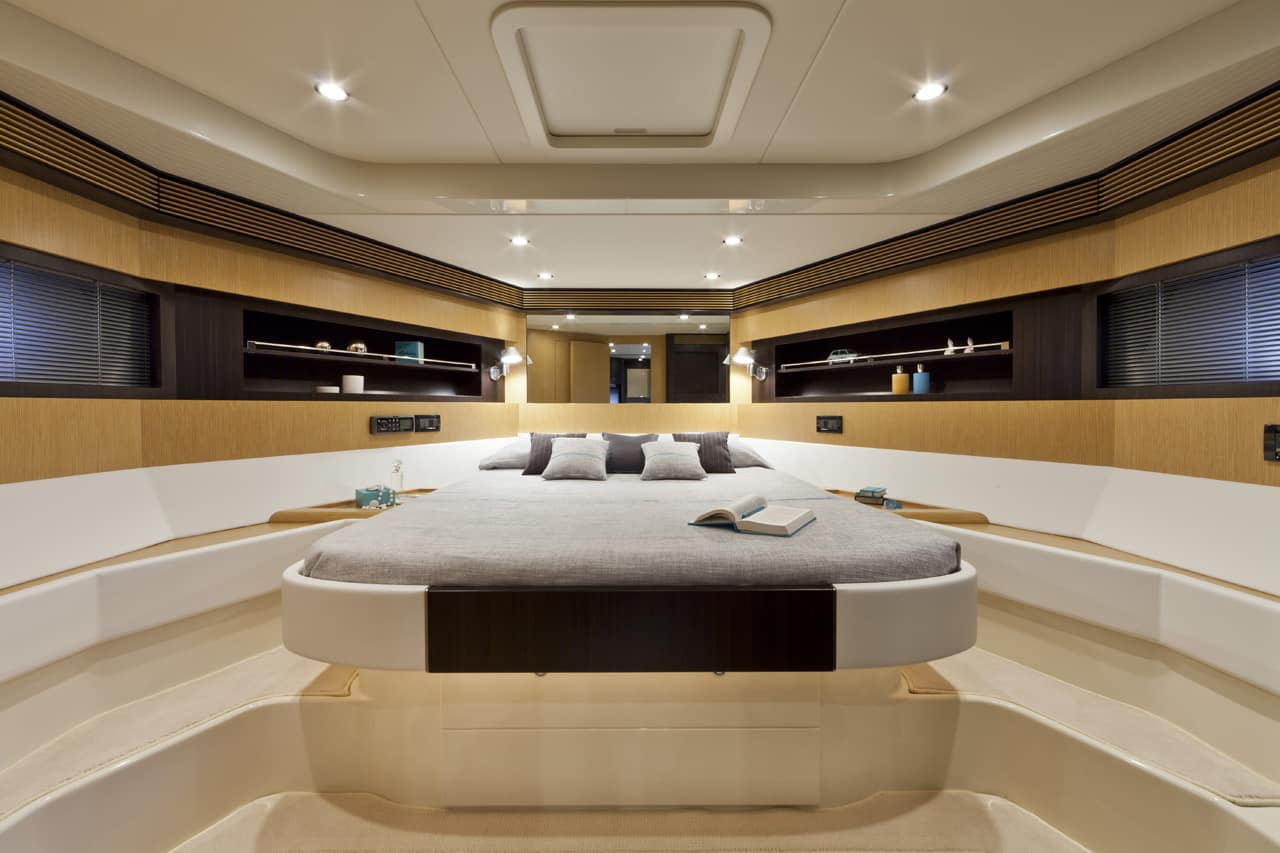 A luxury yacht master bedroom featuring a raised platform bed with gray bedding, surrounded by warm wood paneling and built-in shelves under recessed ceiling lights.