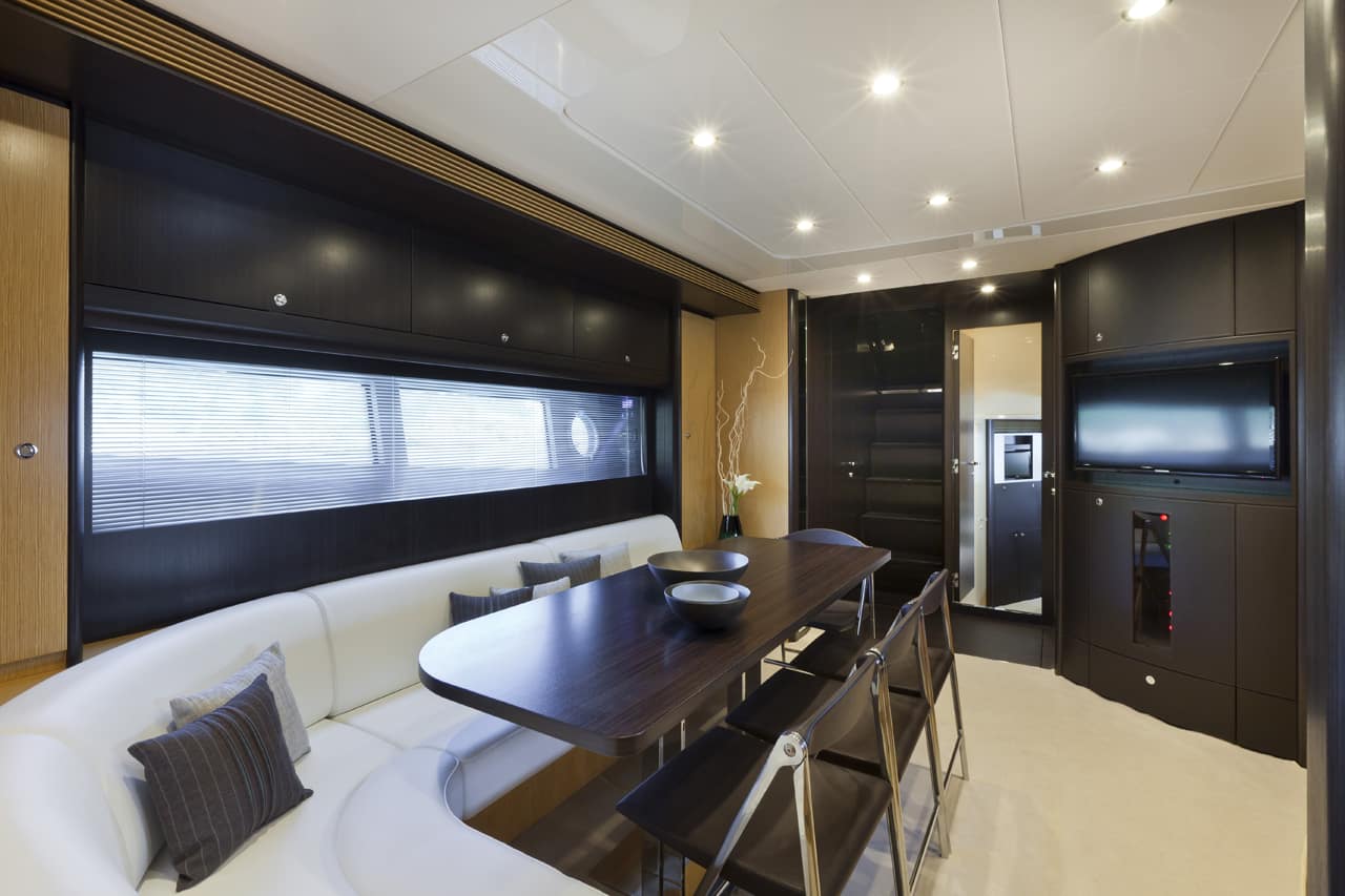 Luxury yacht dining and seating area featuring a curved white leather sofa with dark gray pillows, a dark wood dining table, and modern dark wood cabinetry with a television mounted on the wall.