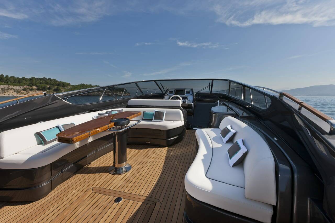 A luxury yacht deck featuring a curved wooden table, white and black upholstered seating, and a wooden teak deck under a blue sky with sea in the background.