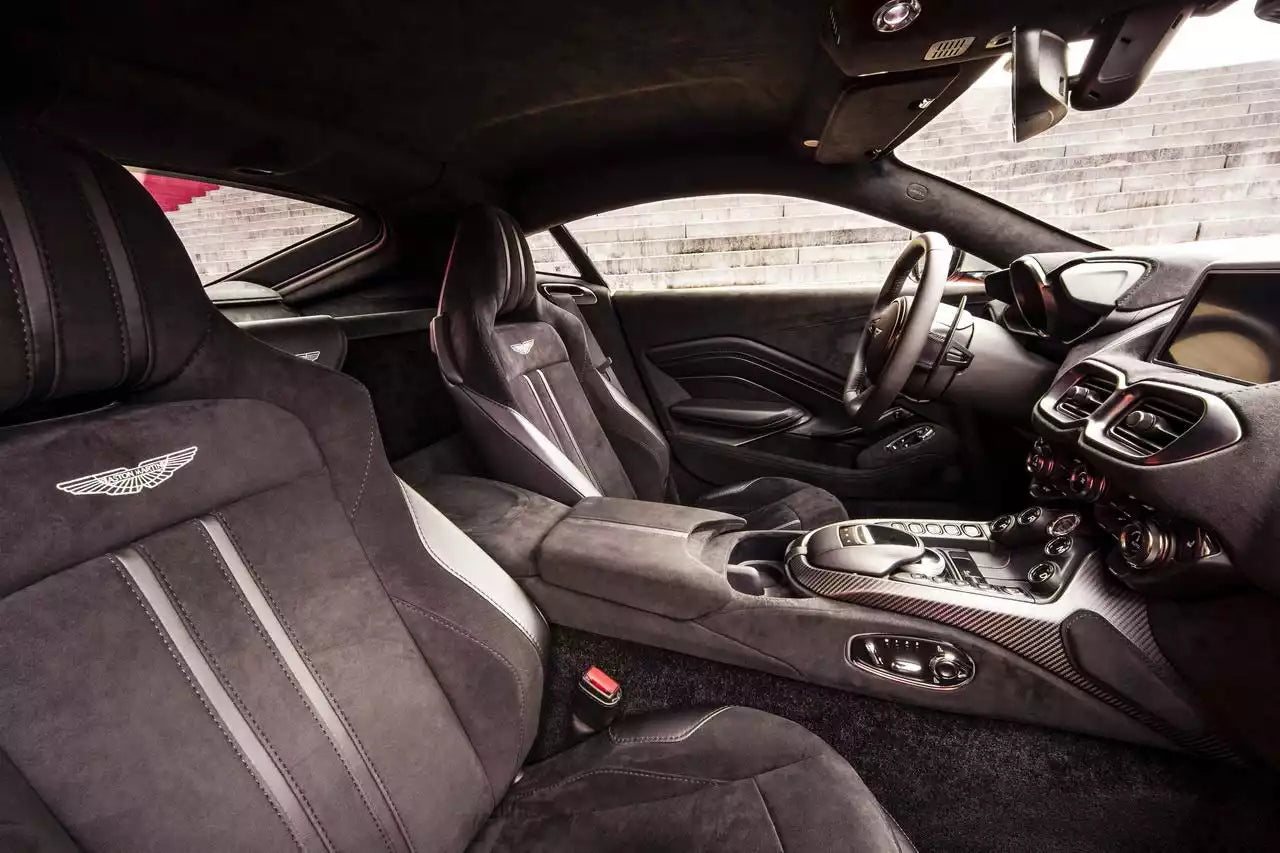 A luxury sports car interior with black and dark gray seats, carbon fiber trim, and visible Aston Martin wing emblems on the front seats.