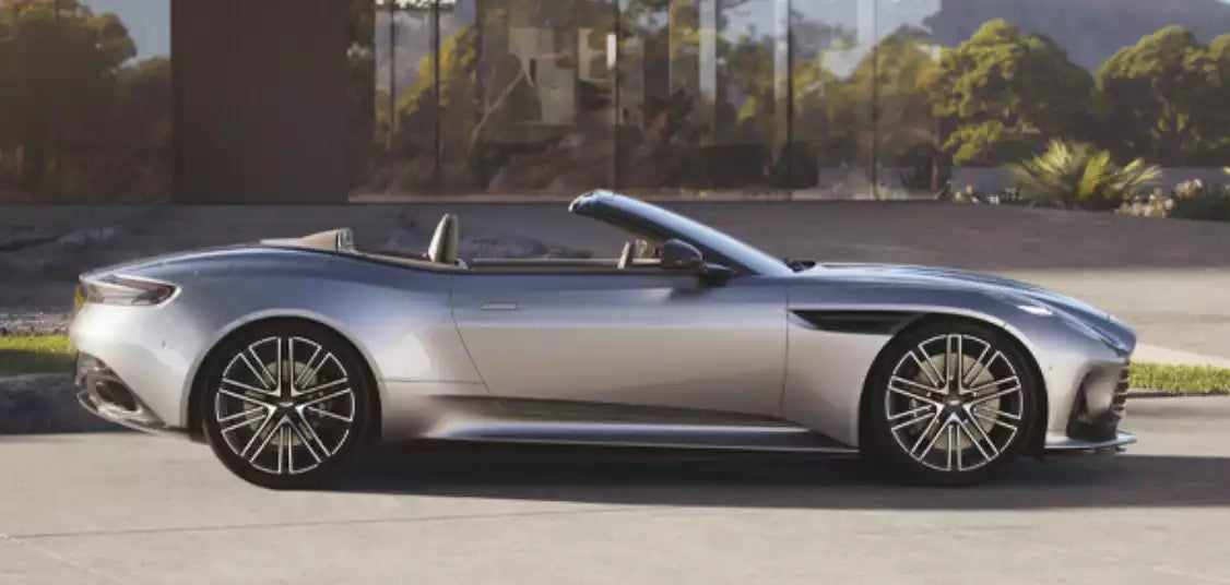 A silver Aston Martin DB10 Volante convertible sports car shown in profile against a modern glass building and outdoor background.