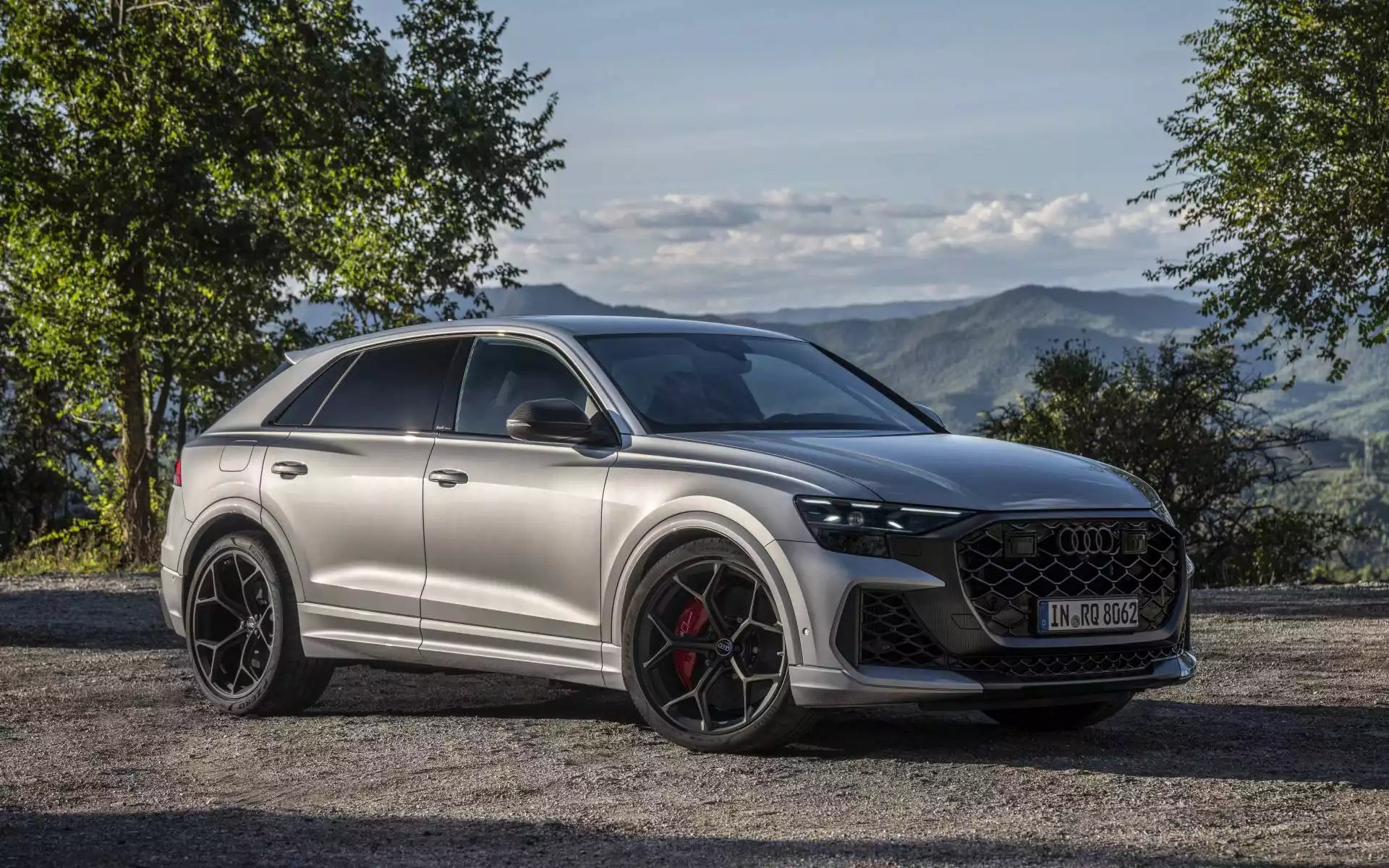 A silver Audi Q2 RS is parked outdoors on a gravel path with a scenic mountain landscape and trees in the background. The car features black multi-spoke wheels with red brake calipers and a large black honeycomb grille.
