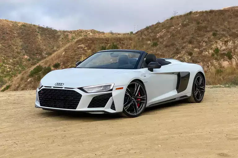 A white Audi R8 Spyder convertible sports car parked on a dirt road with dry hills in the background.