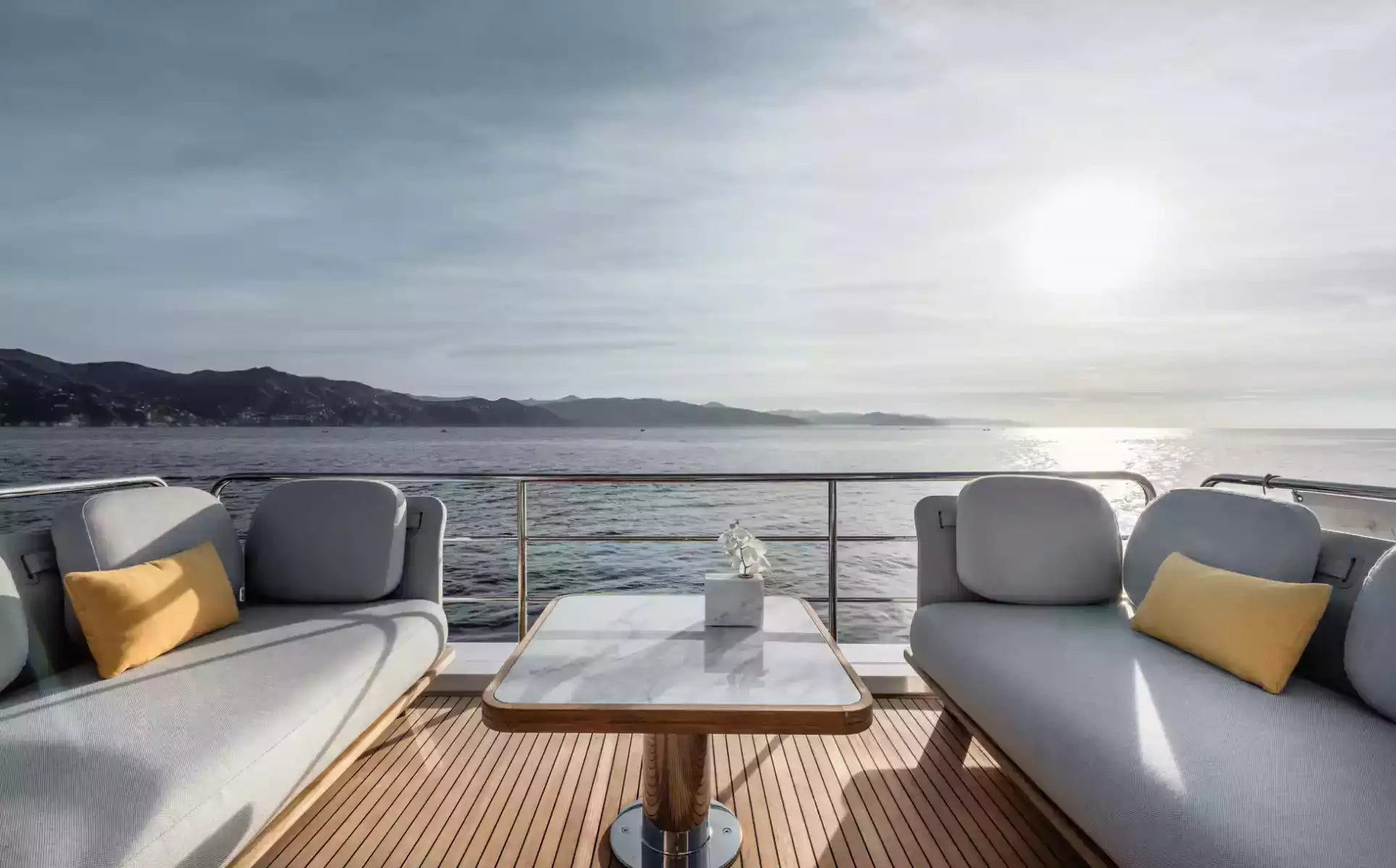 Yacht deck with gray sofas, yellow pillows, and a small table overlooking water and mountains.