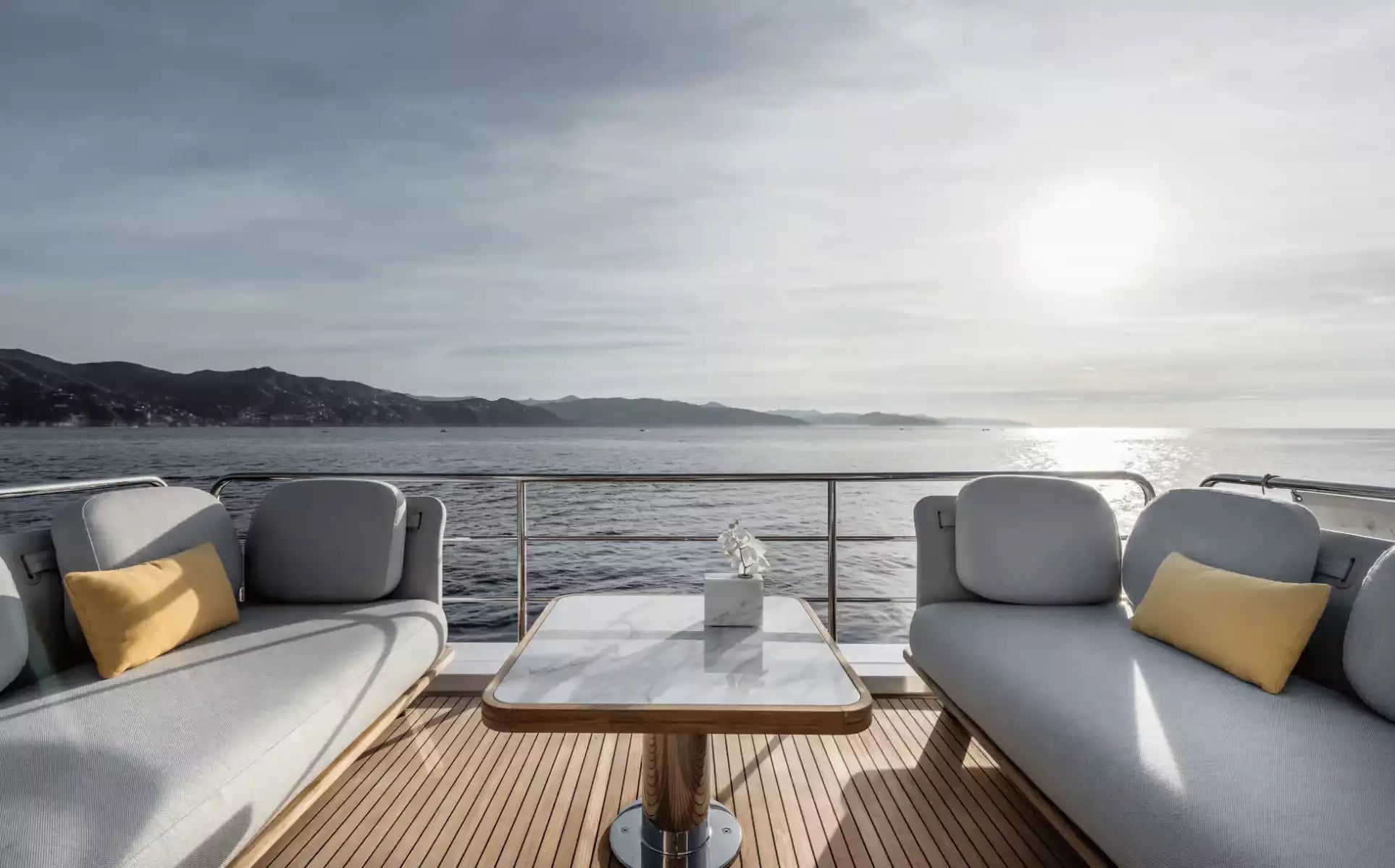 Outdoor seating area on a boat with a table and cushions, overlooking water and mountains.