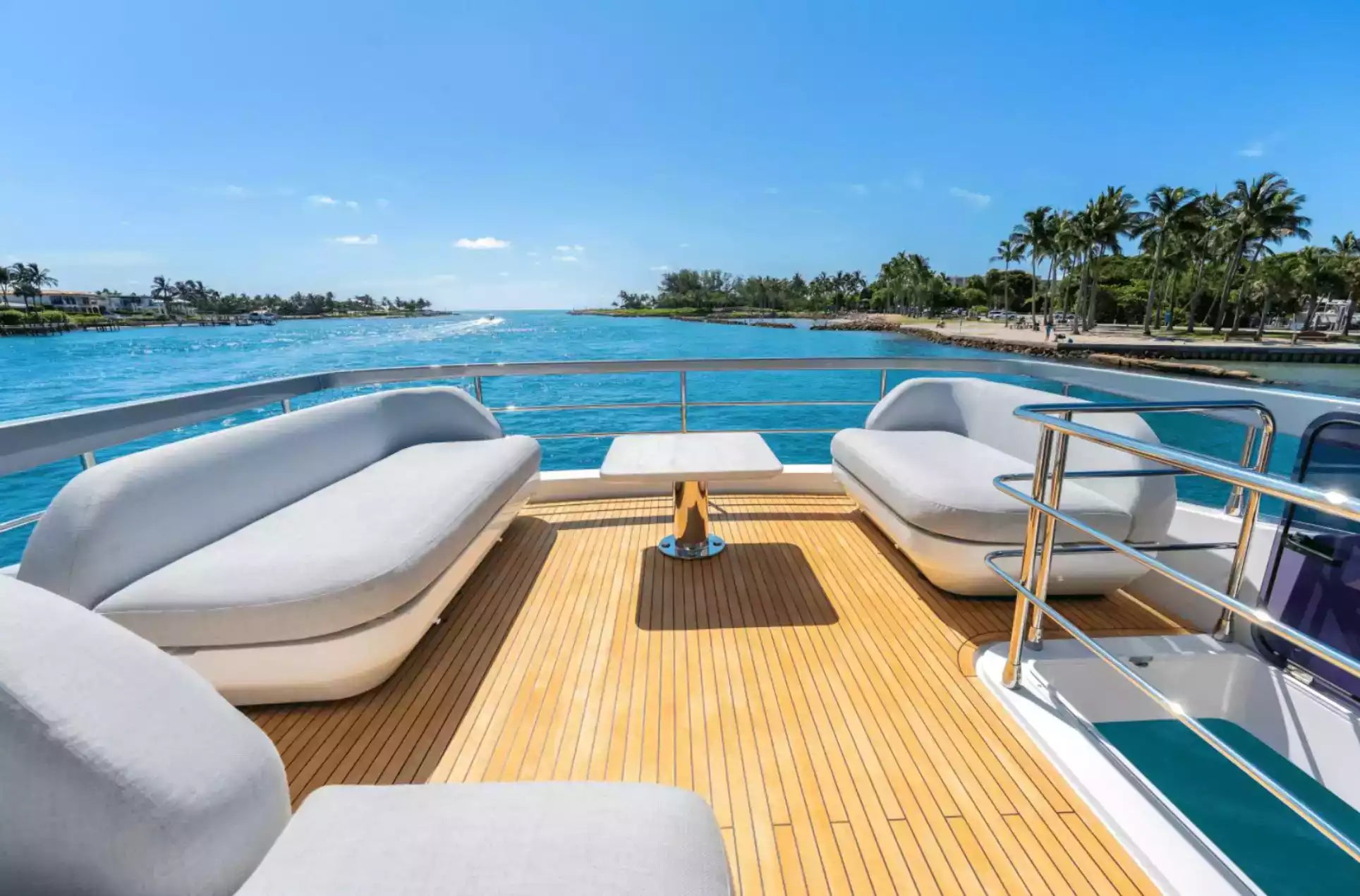A luxury yacht deck featuring light gray cushioned lounge chairs, a small central table, and a wooden teak-style deck floor, overlooking a tropical blue waterway with palm trees under a clear sky.