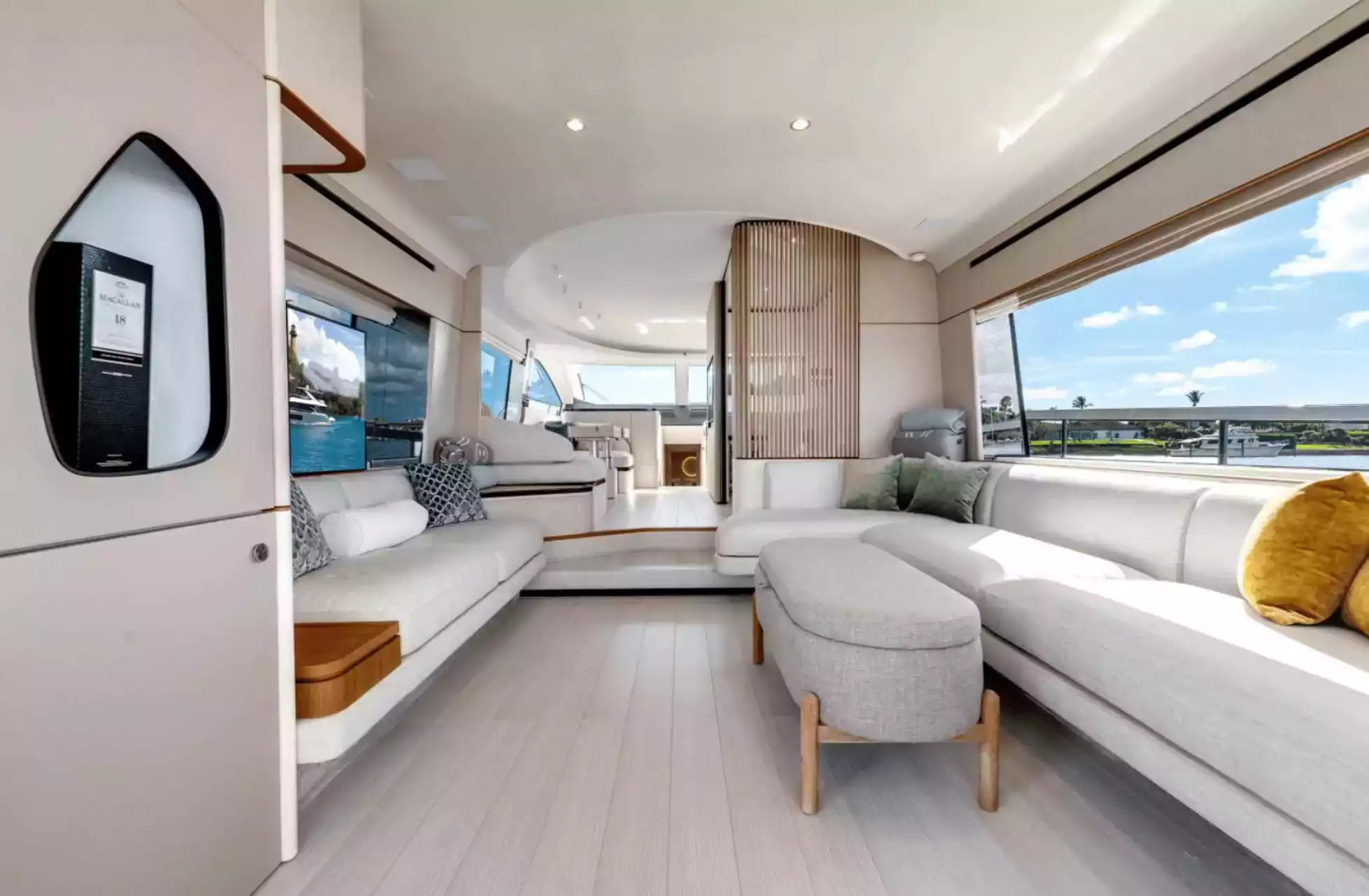 A luxury yacht or modern boat interior with a spacious living area featuring a light gray sectional sofa, matching ottoman, wood flooring, large windows, and a view deck in the background.