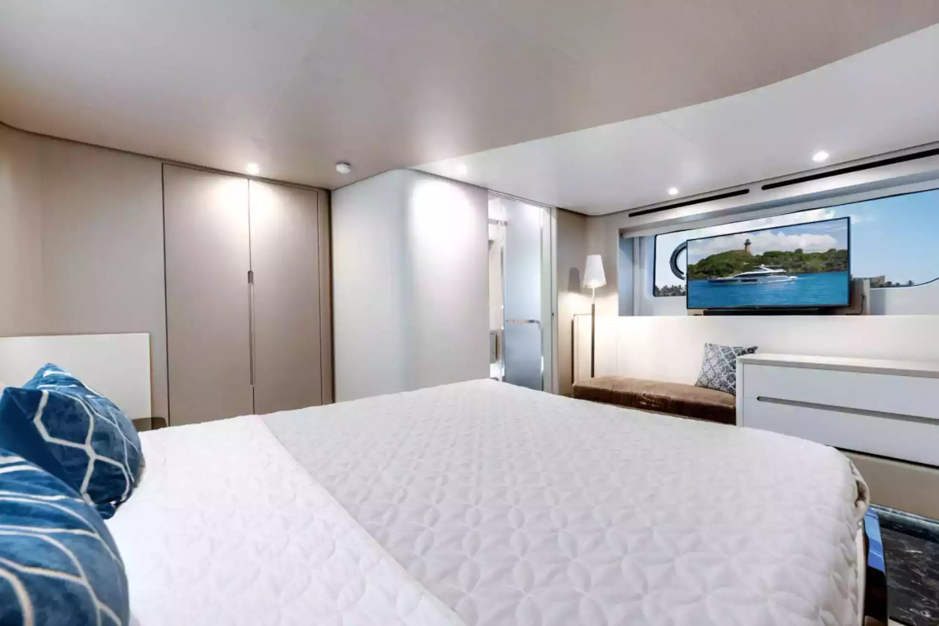 A modern bedroom interior with a large bed in the foreground covered in white bedding, blue patterned pillows, built-in wardrobes, a bench with pillows, and a wall-mounted TV showing a scenic image.