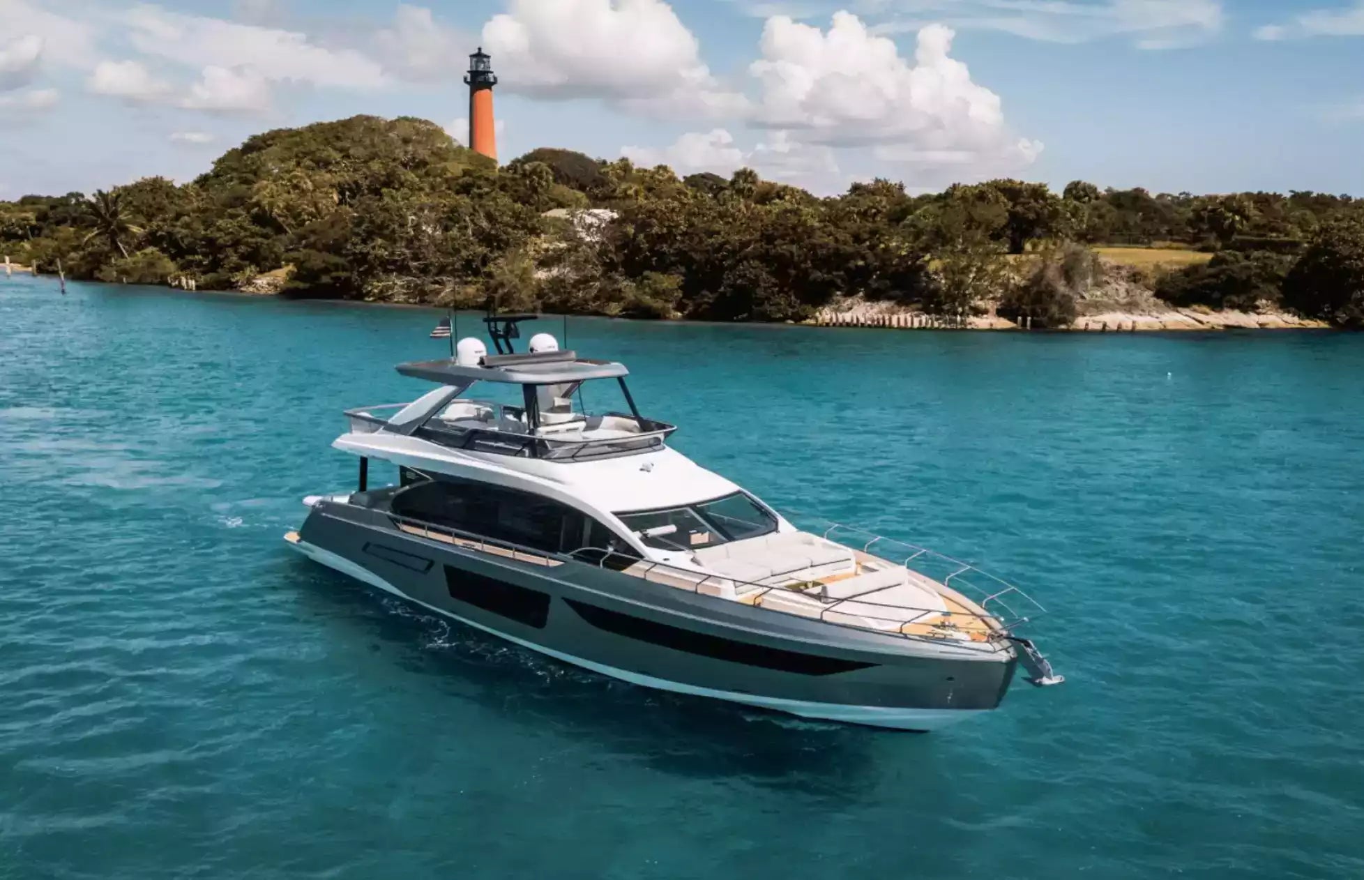 A luxury motor yacht with a gray and white exterior sails on bright turquoise water, with a tropical island featuring a lighthouse and green trees in the background.