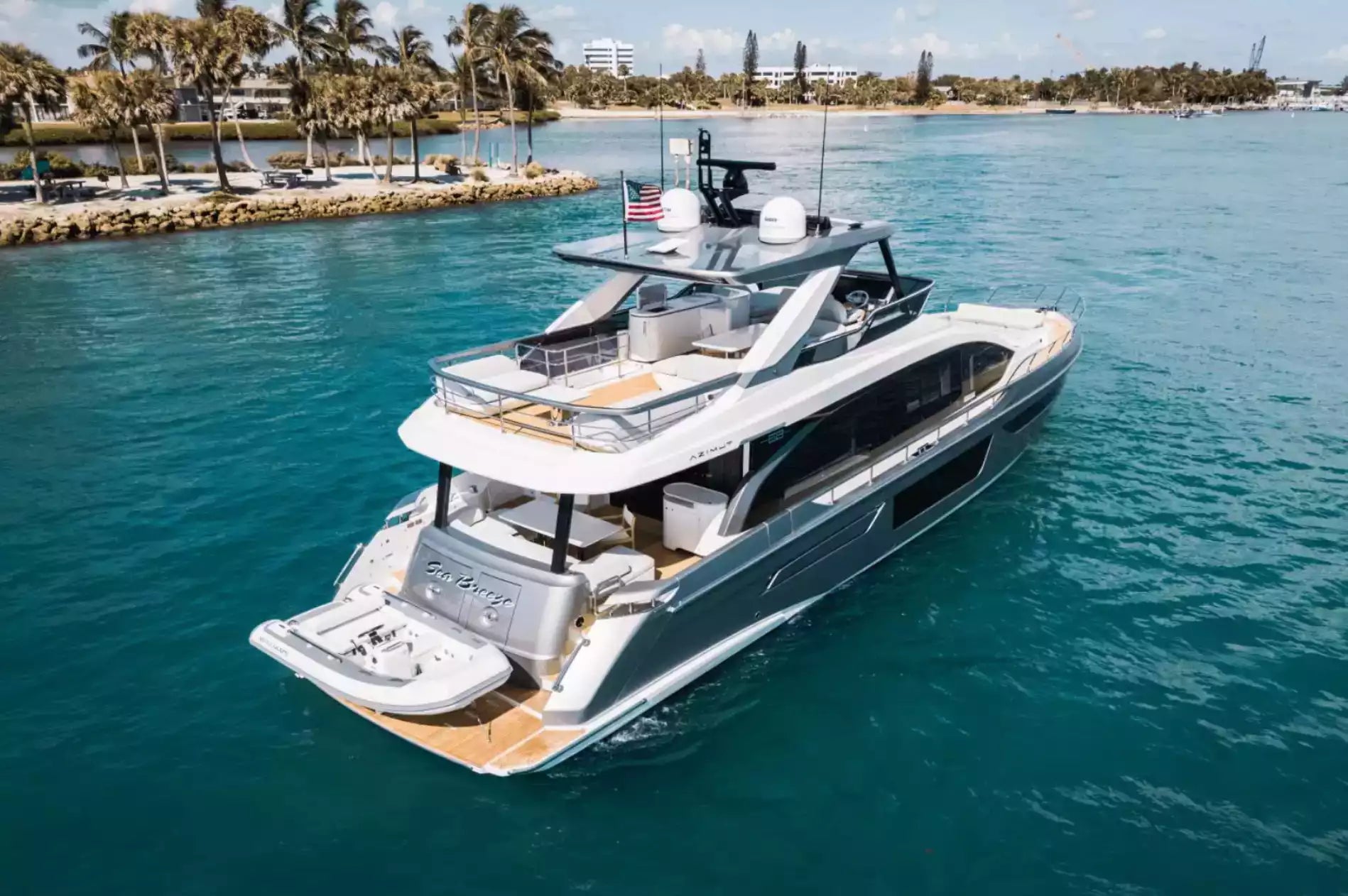 A luxury motor yacht with a tender boat attached, sailing on turquoise water near a tropical shoreline with palm trees and a boardwalk.
