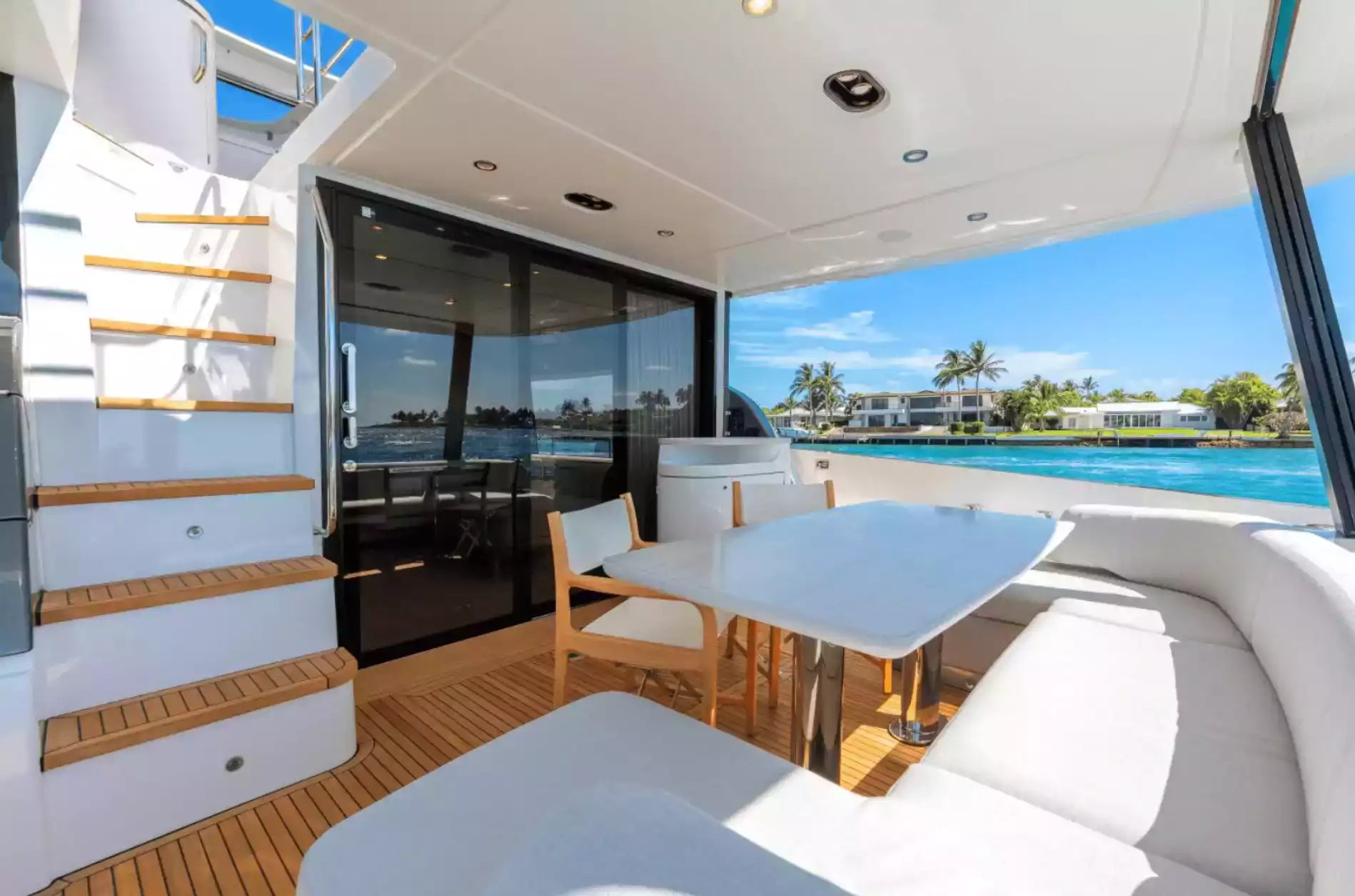 A luxury yacht dining area with a white table and chairs, white seating, teak wood flooring, and large windows overlooking a tropical blue water scene.