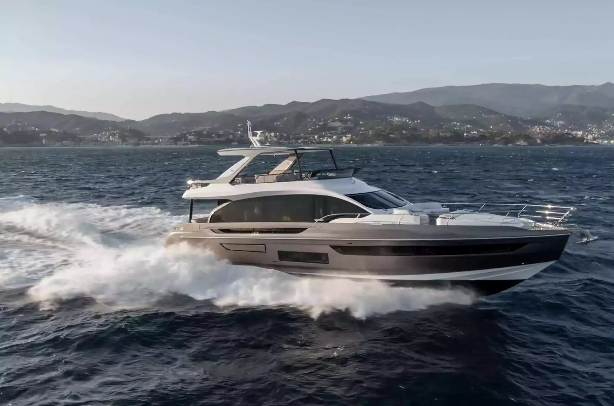 A luxury motor yacht with a dark gray and white hull speeds across deep blue water, creating a white wake, with a hazy coastal mountain range in the background.