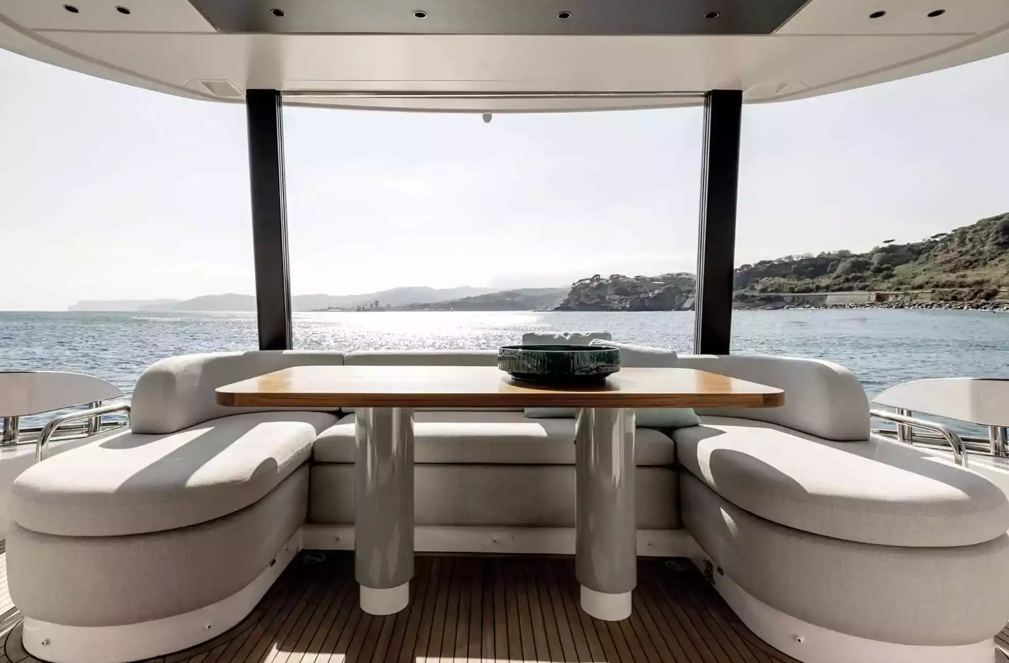 A luxury yacht dining area featuring a round wooden table with metallic legs, flanked by curved white upholstered seating, overlooking a scenic sea and coastline.