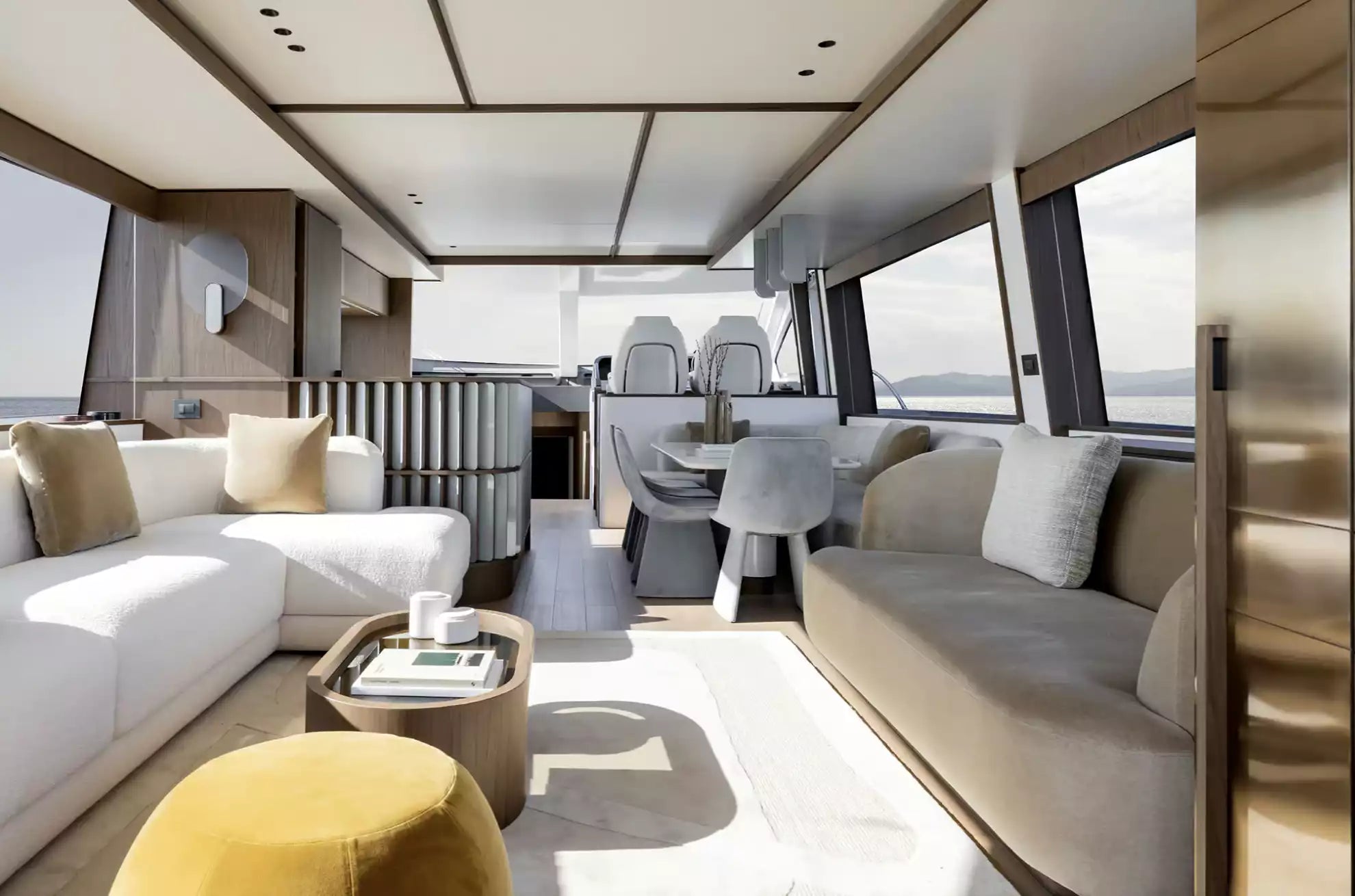 Luxury yacht interior featuring a modern living area with white and beige sofas, a round wooden coffee table, a mustard yellow ottoman, and large windows overlooking the open sea.
