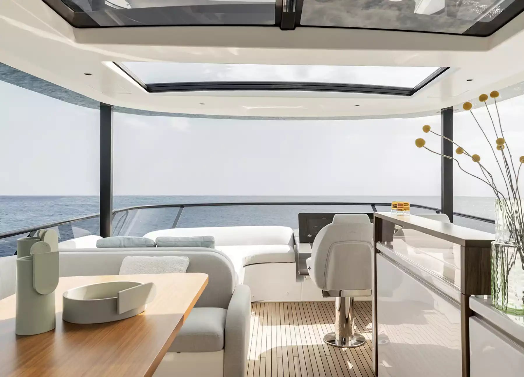 Modern yacht saloon interior with a wooden dining table, white upholstered seating, a high bar counter, and large windows overlooking the ocean.
