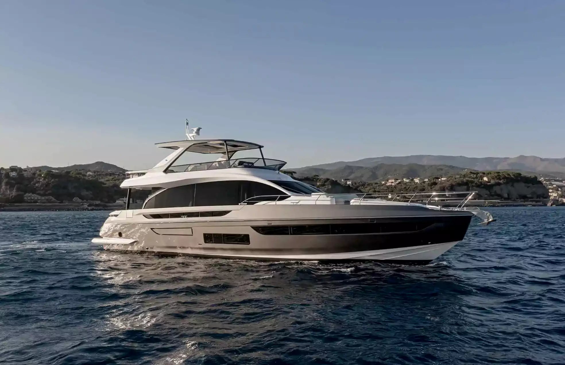 A luxury motor yacht with a white upper deck and a dark gray hull, sailing on deep blue water with a coastal landscape and mountains in the background under a clear blue sky.