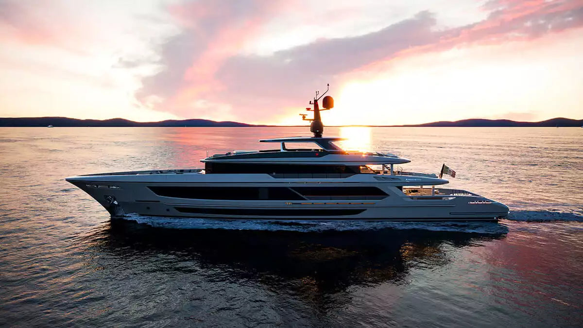 A luxury yacht sailing on open water during a sunset, with pink and orange clouds reflected on the water and a dark shoreline in the distance.