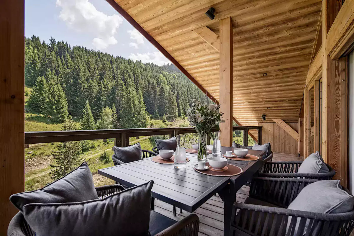 A wooden dining table and matching chairs with gray cushions are set on a balcony overlooking a lush green forest valley.
