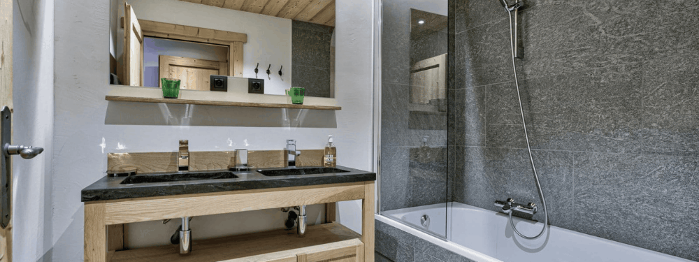 A modern bathroom interior featuring a double-sink vanity with a dark countertop and light wood cabinet, a large mirror, and a glass-enclosed shower area next to a white bathtub.