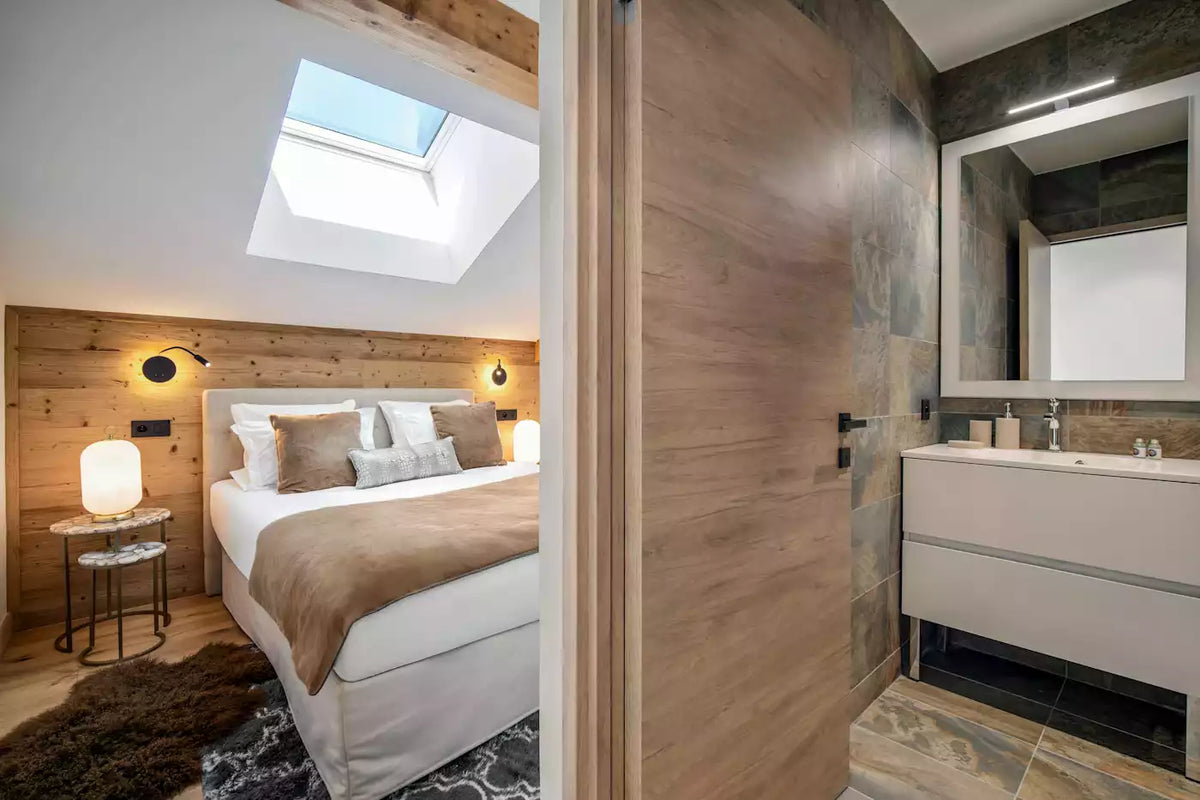 A modern bedroom and bathroom interior with a light wood sliding door separating the spaces. The bedroom features a bed with white linens and a brown throw, wood-paneled walls, bedside tables, and a skylight above.