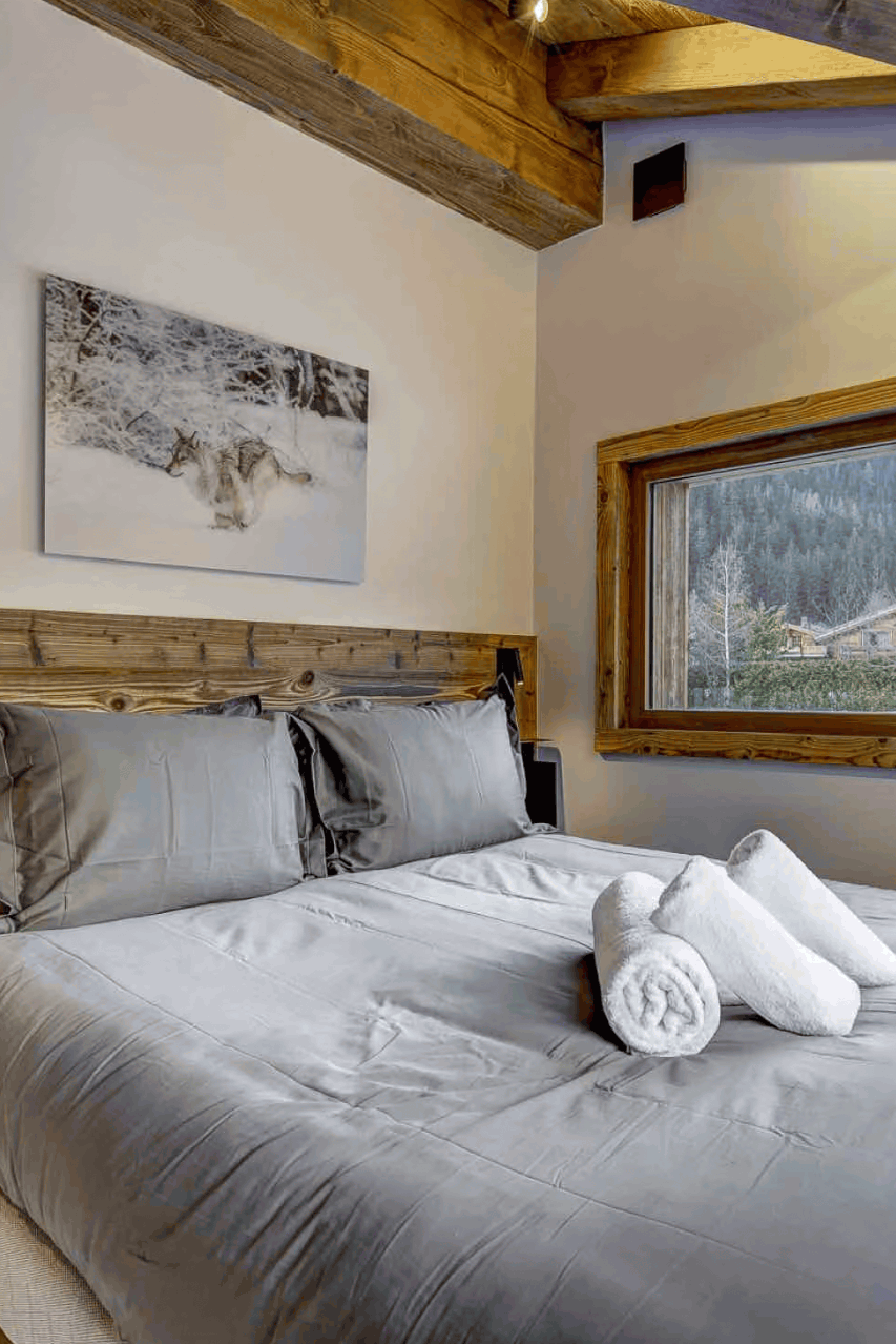 A modern bedroom featuring a bed with gray bedding, a rustic wooden headboard and ceiling beams, and a large window showing a mountain landscape outside.