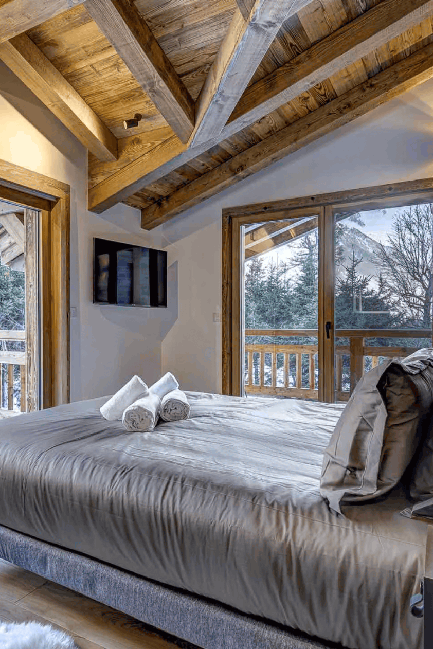 A modern mountain chalet bedroom featuring a large bed with gray bedding, rustic wooden ceiling beams, and floor-to-ceiling windows overlooking a snowy mountain landscape.