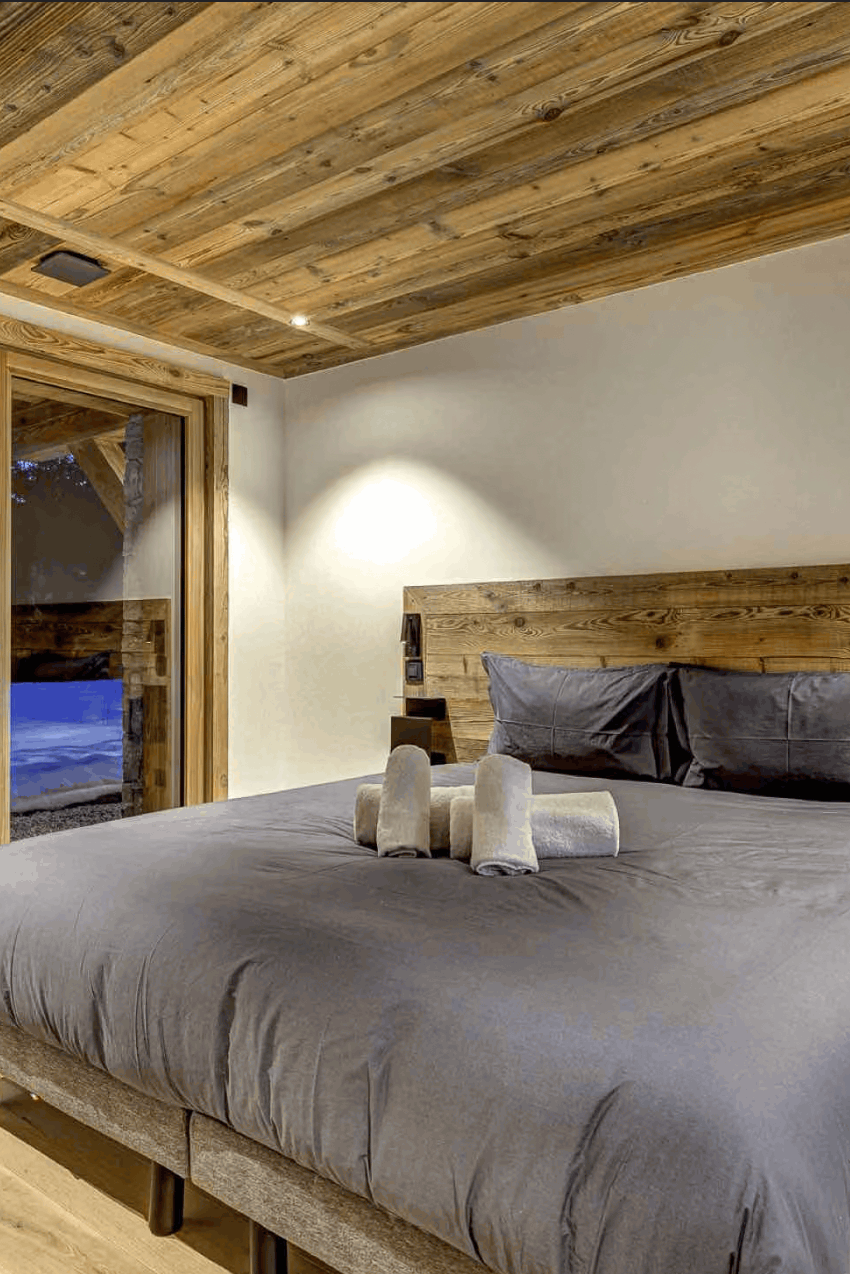 A modern bedroom featuring a large bed with gray bedding, a rustic wooden headboard and ceiling, and floor-to-ceiling glass doors looking out onto a snowy outdoor scene.