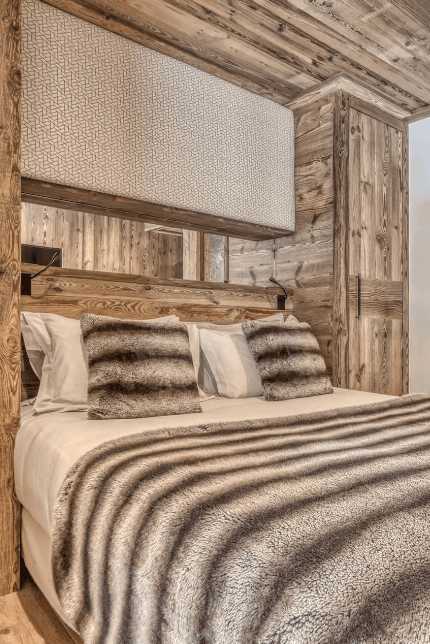A rustic bedroom interior with a large bed dressed in white bedding and a brown-and-white striped animal-print throw blanket, surrounded by wood-paneled walls and ceiling. A doorway to a stone bathroom is visible on the right.