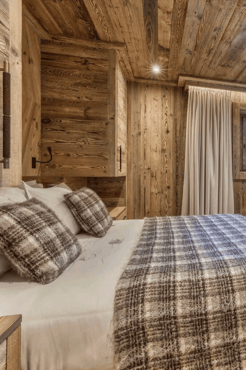 A cozy bedroom with rustic wooden walls and ceiling, featuring a bed dressed in white linens and a brown-and-gray plaid blanket, matching pillows, a small desk by a window, and warm ambient lighting.