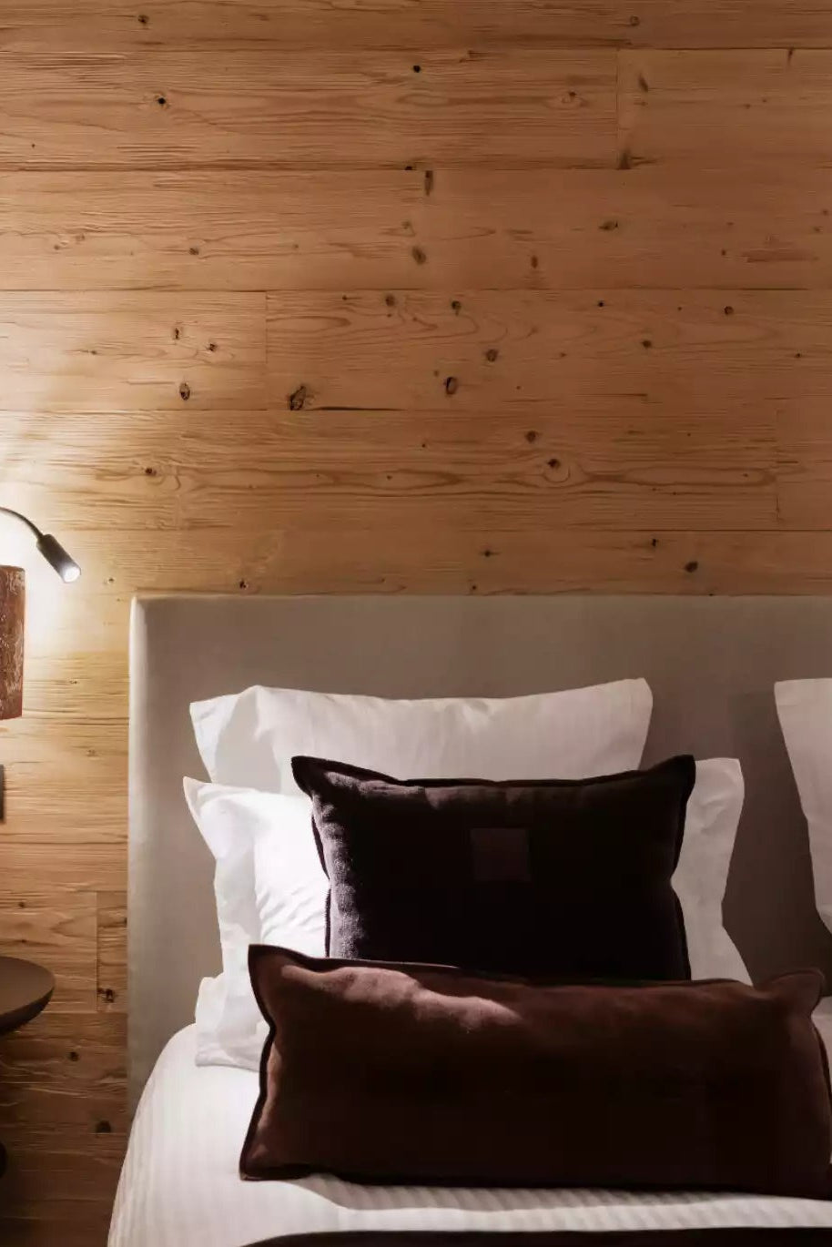 A cozy bedroom scene with a neatly made bed featuring white and dark brown pillows, set against a light wood-paneled wall with a bedside table and lamp.