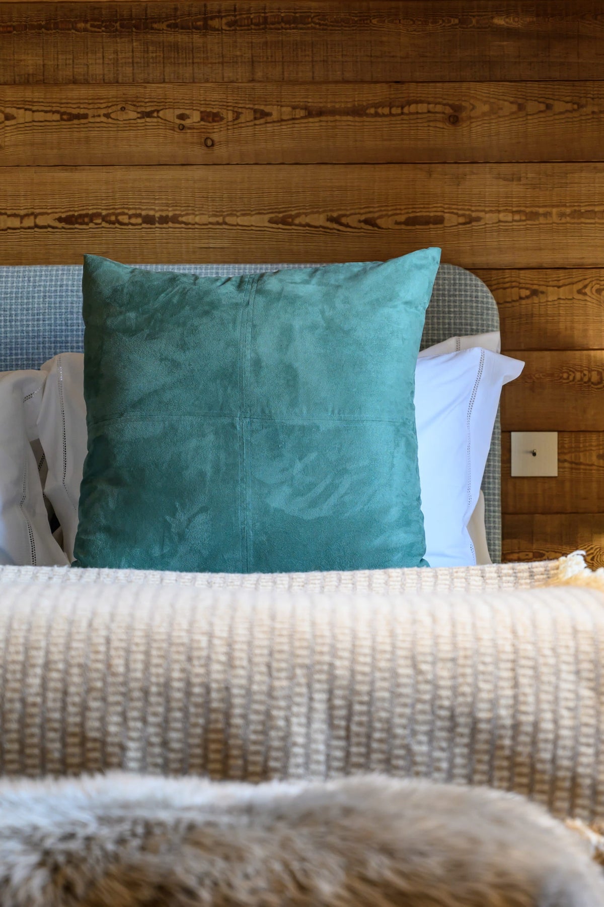 A close-up of a bed styled in a rustic bedroom, featuring two prominent teal velvet pillows, a beige textured knit blanket, white pillows, and a warm wood-paneled wall background.