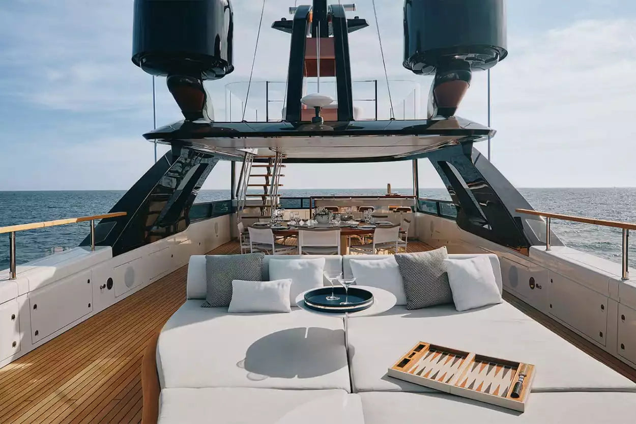 A luxury yacht deck featuring a large white outdoor lounge area with gray and white pillows, a small round table, a wooden tray, and an open view of the ocean and sky.