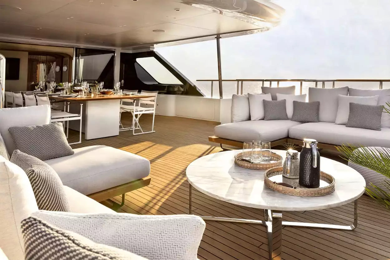 Luxury outdoor patio furniture set on a yacht deck, featuring a modern curved lounge chair, a matching sectional sofa with gray pillows, a round white marble coffee table, and a dining area in the background.