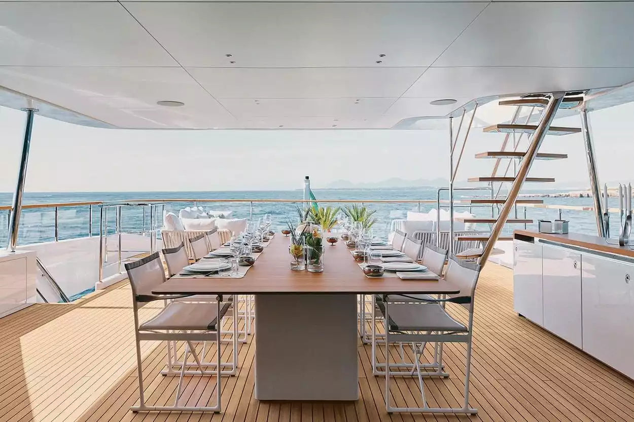 A luxury yacht dining area featuring a long wooden table set with white tableware and glass chairs, overlooking the blue ocean under a bright sky.