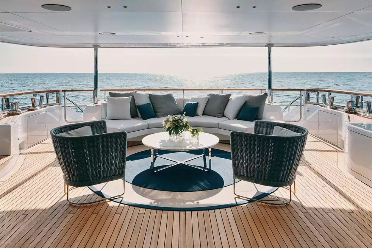 Luxury yacht outdoor lounge area featuring a curved white sofa with gray and navy cushions, two dark woven armchairs, a round white coffee table, and a dark blue circular rug on a wooden deck with the ocean in the background.